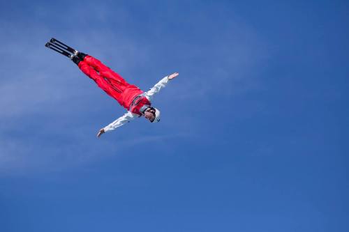 A skier performs a mid-air trick against a clear blue sky, arms outstretched and wearing a red outfit.