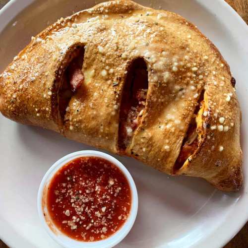 A golden-brown calzone with visible fillings, served with a side of marinara sauce and grated cheese.