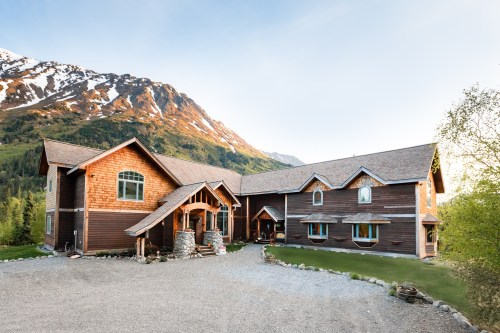 A large wooden house with a stone entrance, surrounded by mountains and greenery, under a clear blue sky.