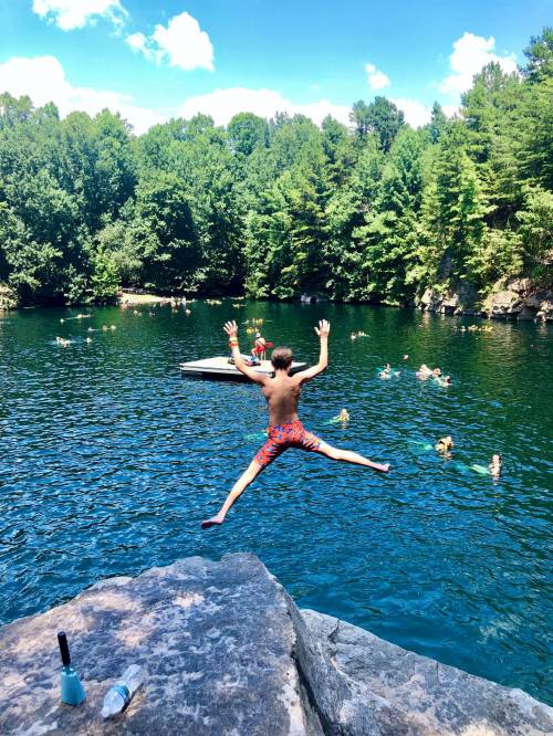 A person jumps off a rock into a clear lake surrounded by trees, with others swimming nearby on a sunny day.