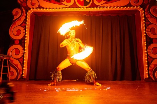 A performer in tribal attire spins flaming torches on stage, surrounded by vibrant decorations and a dark backdrop.
