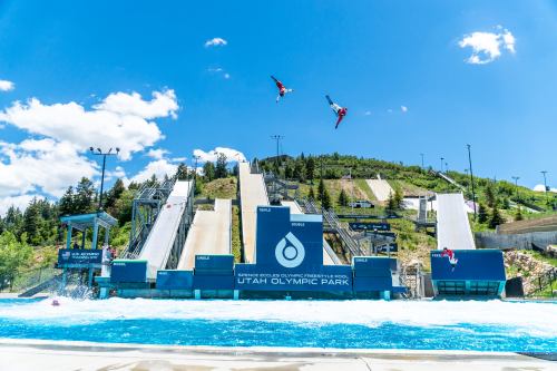 Two athletes perform aerial tricks above a water ramp at Utah Olympic Park, with ski jumps and a blue sky in the background.