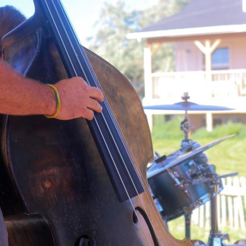A close-up of a hand playing a double bass, with a drum set visible in the background and a house in the distance.