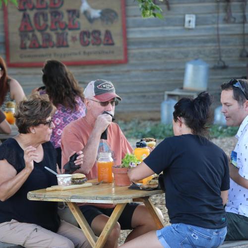 A group of people enjoying food and drinks at a rustic outdoor table at a farm.