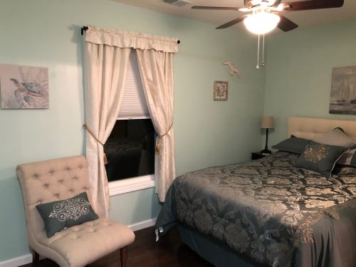 Cozy bedroom with a bed, decorative pillows, a chair, and light blue walls, featuring curtains and wall art.