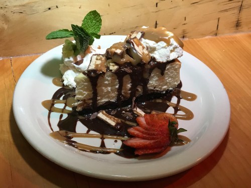 A slice of chocolate cake topped with whipped cream, caramel drizzle, nuts, and a fresh strawberry on a white plate.