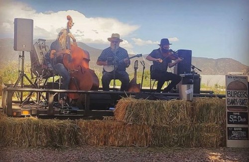 A trio of musicians performs on a trailer surrounded by hay bales, with mountains in the background and a sign for burgers.