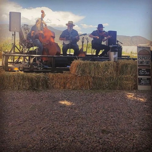 A trio of musicians performs on a stage made of hay bales, with mountains in the background and a sign for burgers nearby.