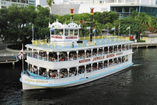 A large, colorful riverboat named "Jungle Queen" cruises along a waterway, filled with passengers enjoying the view.