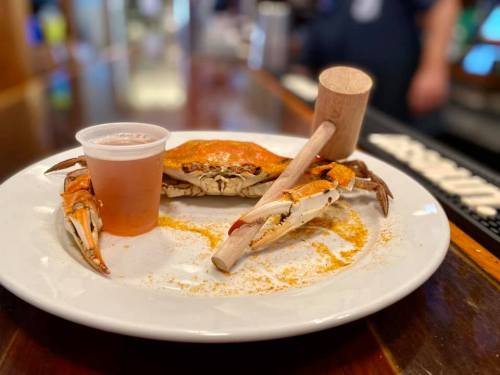 A whole crab on a plate with a wooden mallet and a small cup of beer beside it, set on a wooden table.