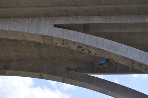 A climber ascends the underside of a concrete bridge, showcasing impressive skill against a blue sky.