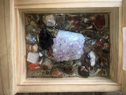 A wooden box filled with various colorful crystals and stones, including a prominent amethyst cluster on top.