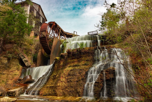 A scenic view of a watermill with a large wheel, cascading waterfalls, and lush greenery surrounding the area.