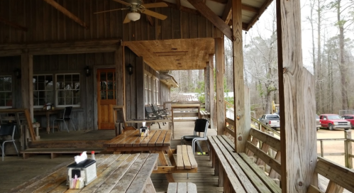 A rustic wooden porch with tables and chairs, overlooking a quiet outdoor area with parked cars in the background.