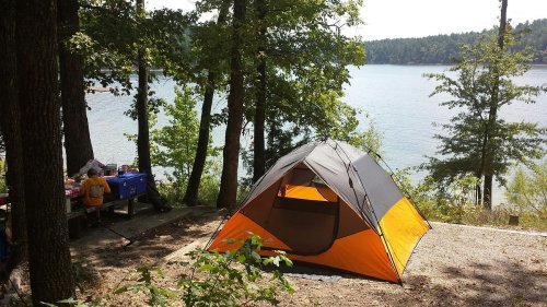 A tent set up by a lake, surrounded by trees, with a picnic table nearby. Bright sunlight illuminates the scene.