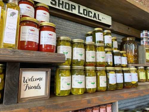 Shelves filled with jars of sauces and oils, with a sign reading "Shop Local" and a welcome sign for visitors.