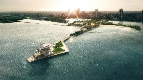 Aerial view of a modern waterfront development with a pier, greenery, and city skyline in the background at sunset.