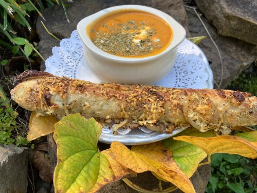 A plate with a crispy bread roll and a bowl of orange soup, garnished with herbs, set on a stone surface with leaves.