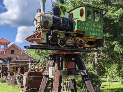 A vintage steam locomotive model on display, mounted on a wooden stand, surrounded by greenery and buildings.
