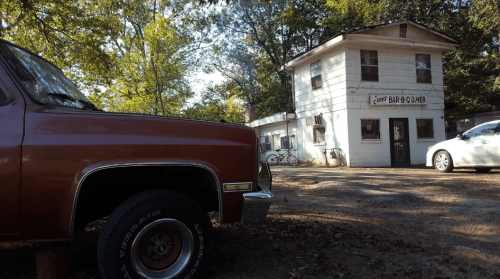 A vintage truck in the foreground with a two-story building labeled "Bar & Grill" in a wooded area.