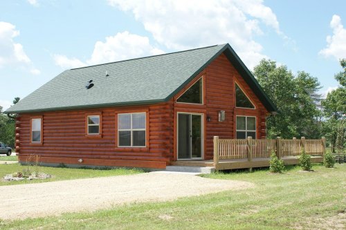 A rustic log cabin with a green roof, front porch, and large windows, set in a grassy area under a blue sky.