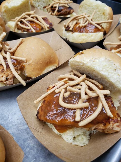 Close-up of chicken sandwiches with barbecue sauce and crispy noodles on soft buns, served in paper trays.