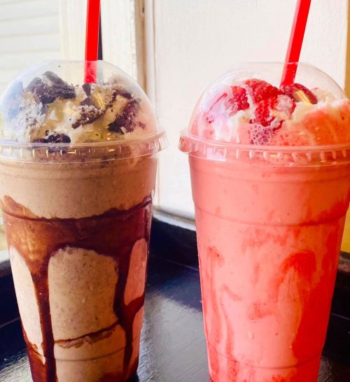 Two colorful milkshakes in clear cups with red straws: one chocolate with cookies, the other strawberry with whipped cream.
