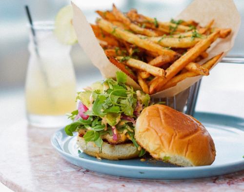 A plate with a colorful burger topped with greens and a side of crispy fries in a basket. A drink is in the background.