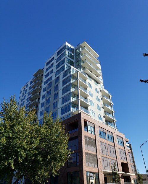 A modern multi-story building with balconies, set against a clear blue sky and surrounded by trees.