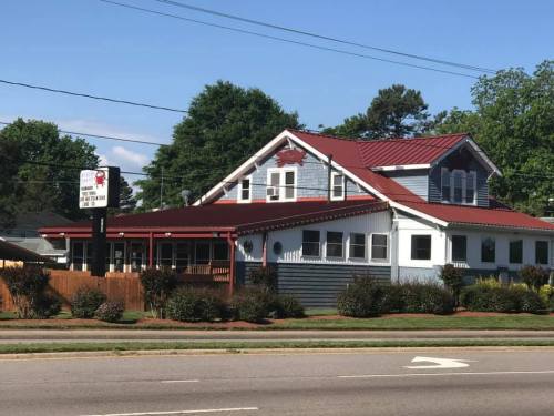 A two-story restaurant with a red metal roof, blue siding, and a sign out front, surrounded by trees and a clear sky.