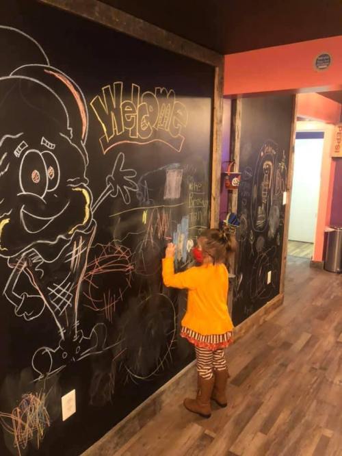A child in an orange outfit draws on a chalkboard wall decorated with colorful chalk art in a playful setting.