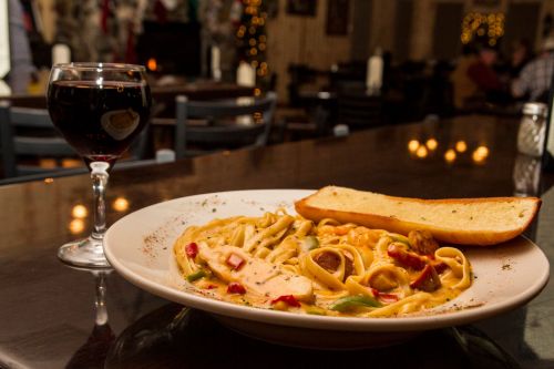 A plate of fettuccine pasta with vegetables and a slice of garlic bread, accompanied by a glass of red wine.