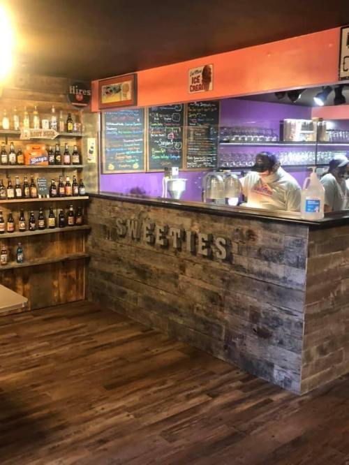 Interior of a cozy ice cream shop named "Sweeties," featuring a colorful menu and shelves of bottled drinks.