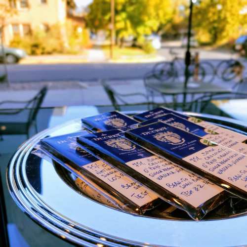 A silver tray holds several chocolate bars with handwritten labels, set against a sunny outdoor scene.