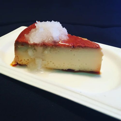 A slice of creamy flan topped with a layer of ice, served on a white plate against a dark background.