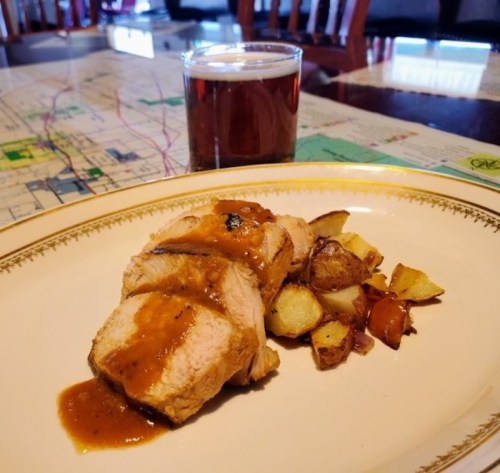 A plate of sliced pork with sauce, roasted potatoes, and a glass of amber beer on a table with a map.