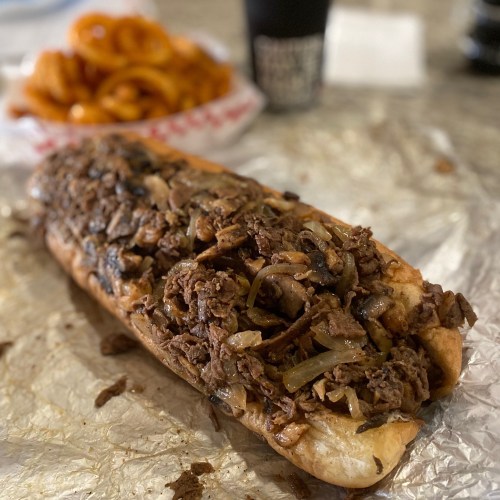 A loaded sandwich filled with beef, onions, and mushrooms, served with a side of curly fries.