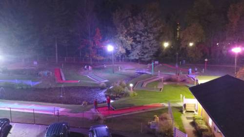 A nighttime view of a mini-golf course with colorful paths, surrounded by trees and illuminated by bright lights.