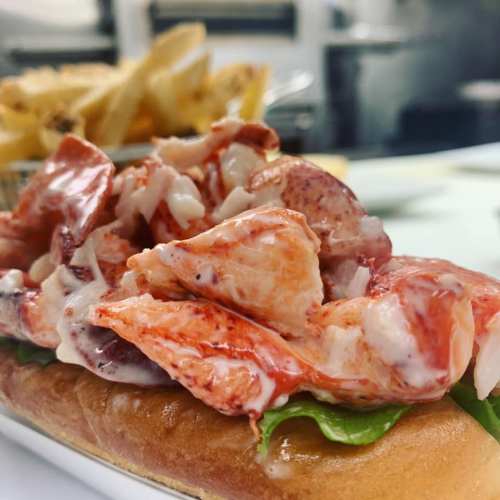 A lobster roll topped with fresh lobster meat and lettuce, served with a side of fries in the background.