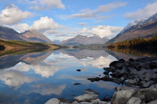 A serene lake reflects mountains and clouds, surrounded by rocky shores and autumn-colored trees.