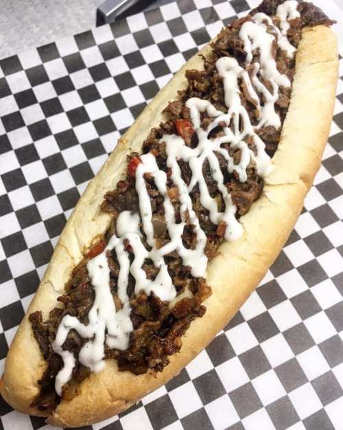 A loaded sandwich on a checkered paper, filled with seasoned meat and drizzled with white sauce.