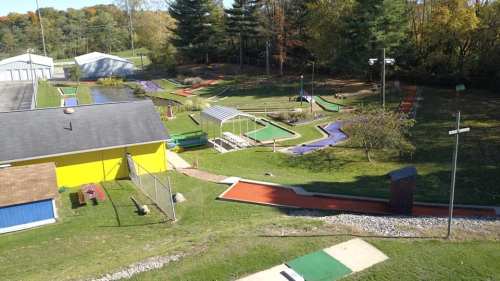 Aerial view of a colorful mini-golf course with various obstacles, surrounded by trees and a small pond.