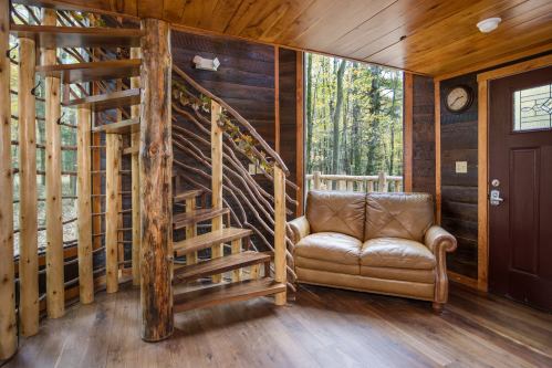 Cozy interior with wooden spiral staircase, leather couch, and large windows overlooking a forested area.