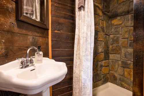 A rustic bathroom featuring a stone shower, white sink, and wooden walls, with a mirror above the sink.