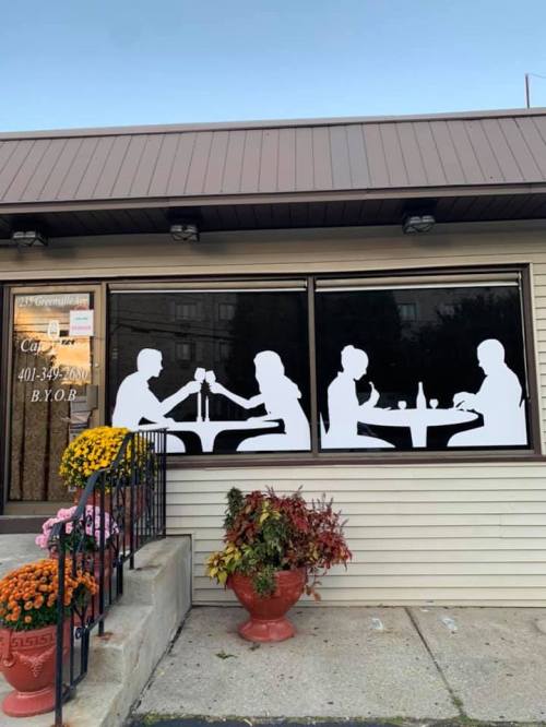 Silhouettes of two couples dining at tables, seen through a restaurant window with flower pots outside.