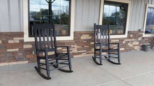 Two black rocking chairs sit on a concrete porch in front of a stone wall and large windows.