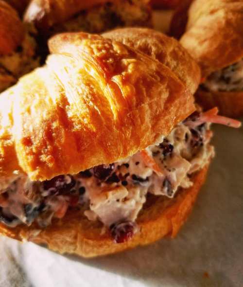 A close-up of a flaky croissant filled with creamy chicken salad, garnished with cranberries and herbs.
