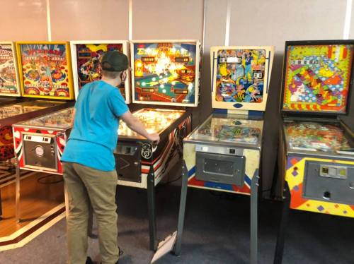 A person in a blue shirt plays pinball at a row of vintage machines in a game room.