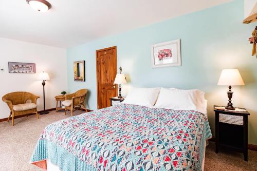 Cozy bedroom with a patterned quilt, two lamps, a small table, and a chair against a light blue wall.