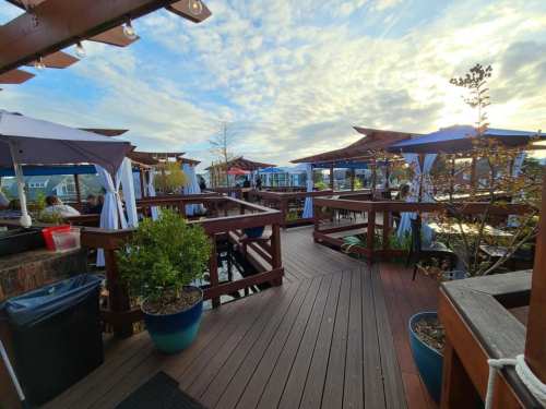 A wooden deck with tables and umbrellas, surrounded by plants, under a cloudy sky at a waterfront restaurant.